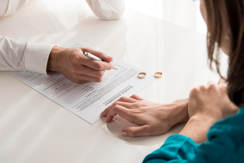 Two people sit at a table as one signs a divorce agreement form, with two wedding rings and divorce financial documents resting on the table.